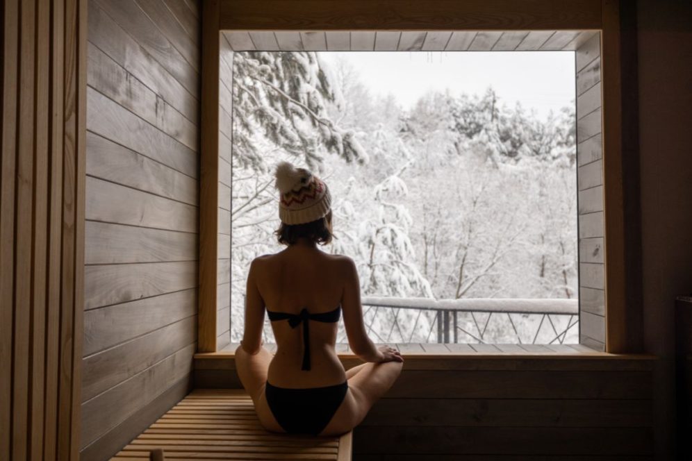 woman in bikini sitting inside sauna, looking out window at snowy winter scenery